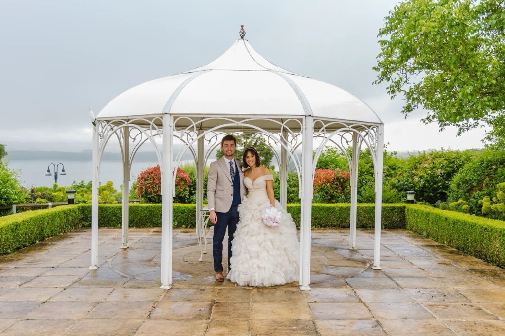 Bride and groom under a gazebo; groom in a beige jacket with navy waistcoat and trousers, hired from Scotney’s of London Road