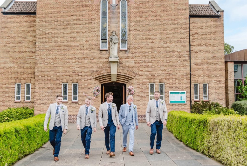 Groomsmen walking outside a church in beige jackets, navy trousers and a light blue suit, hired from Scotney’s of London Road